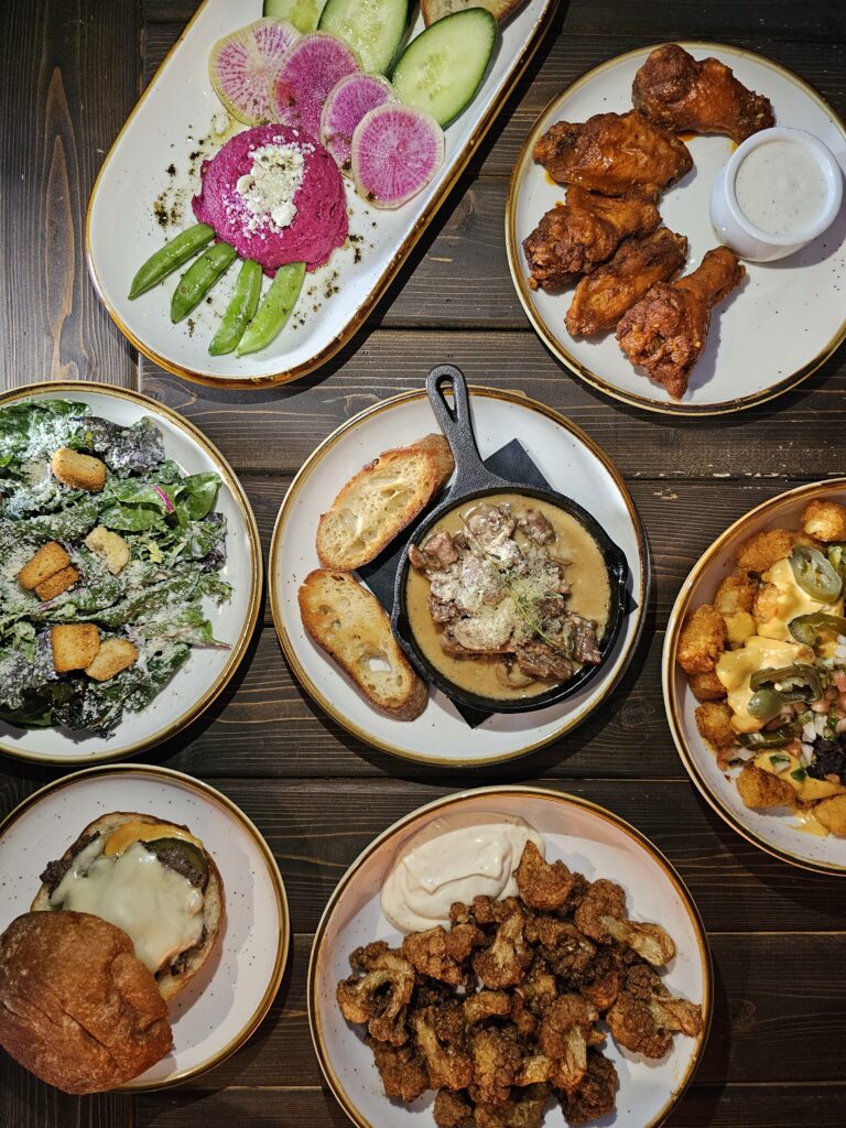 Overhead spread of shareable plates at Camp West — buffalo wings, beet hummus, Caesar salad, loaded tots and a slider