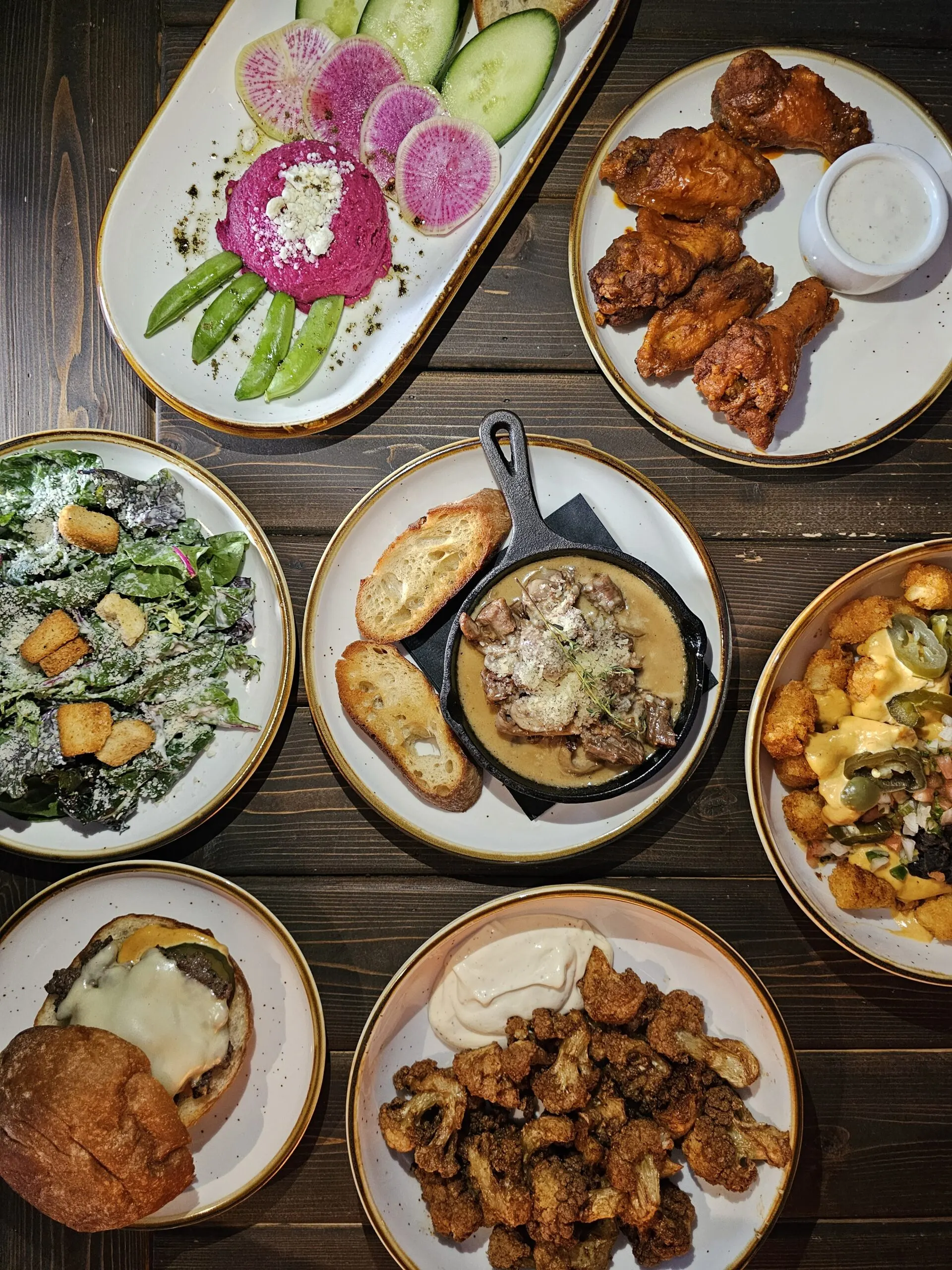 Overhead spread of Camp West shareable plates with beet hummus, wings, a cast iron skillet with toast, kale salad, loaded tots, fried cauliflower and a burger