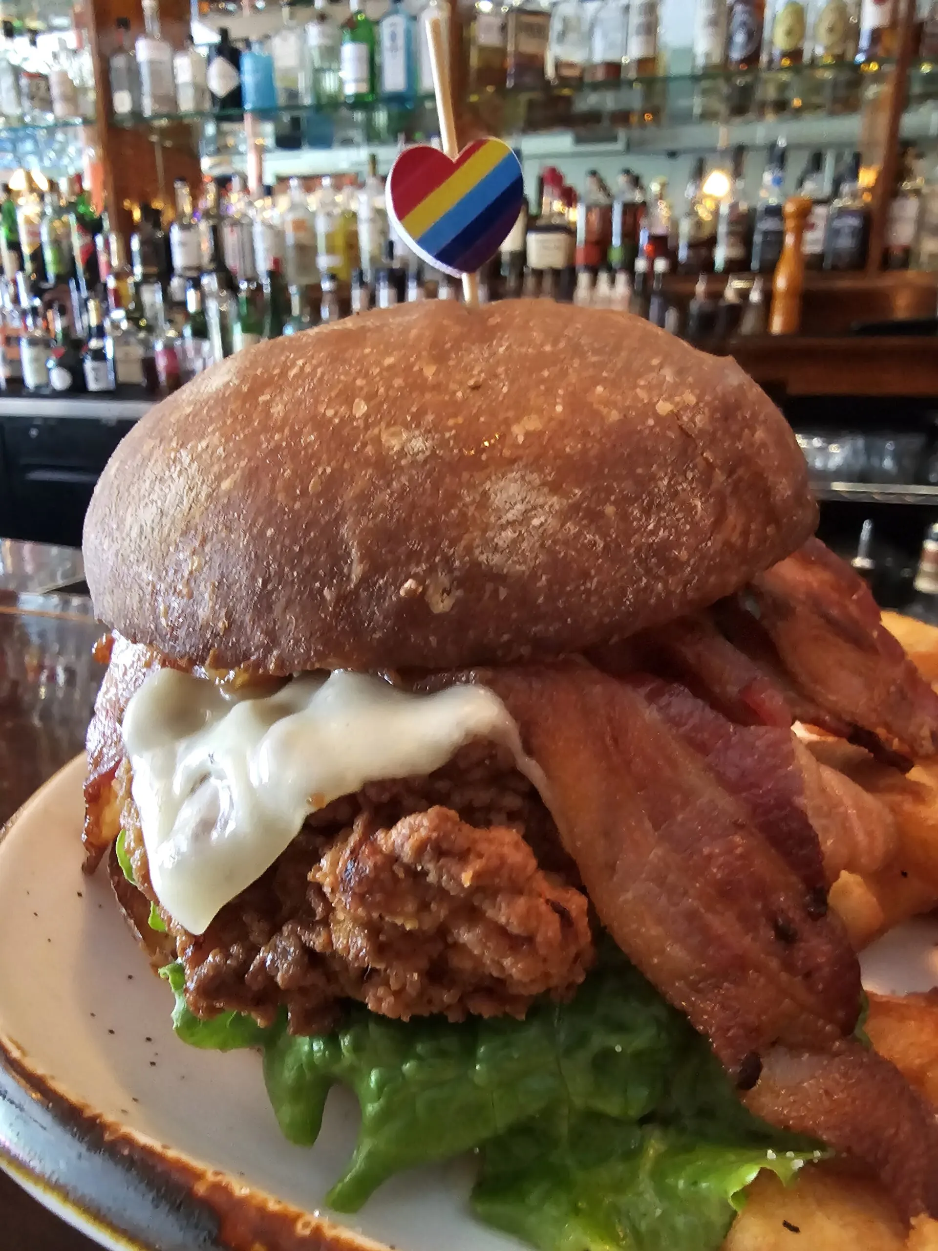 Fried chicken sandwich with bacon and melted cheese on a dark bun at Camp West, with the back bar behind