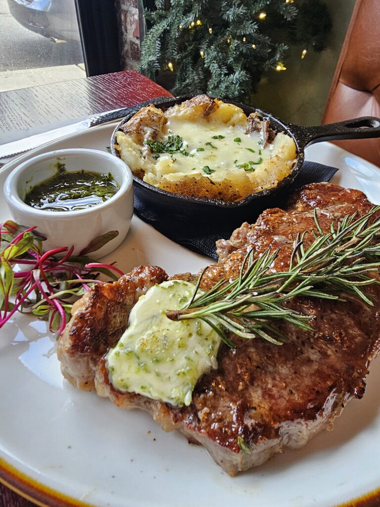 Seared ribeye topped with herb butter and rosemary at Camp West with a cast iron skillet of gratin and chimichurri