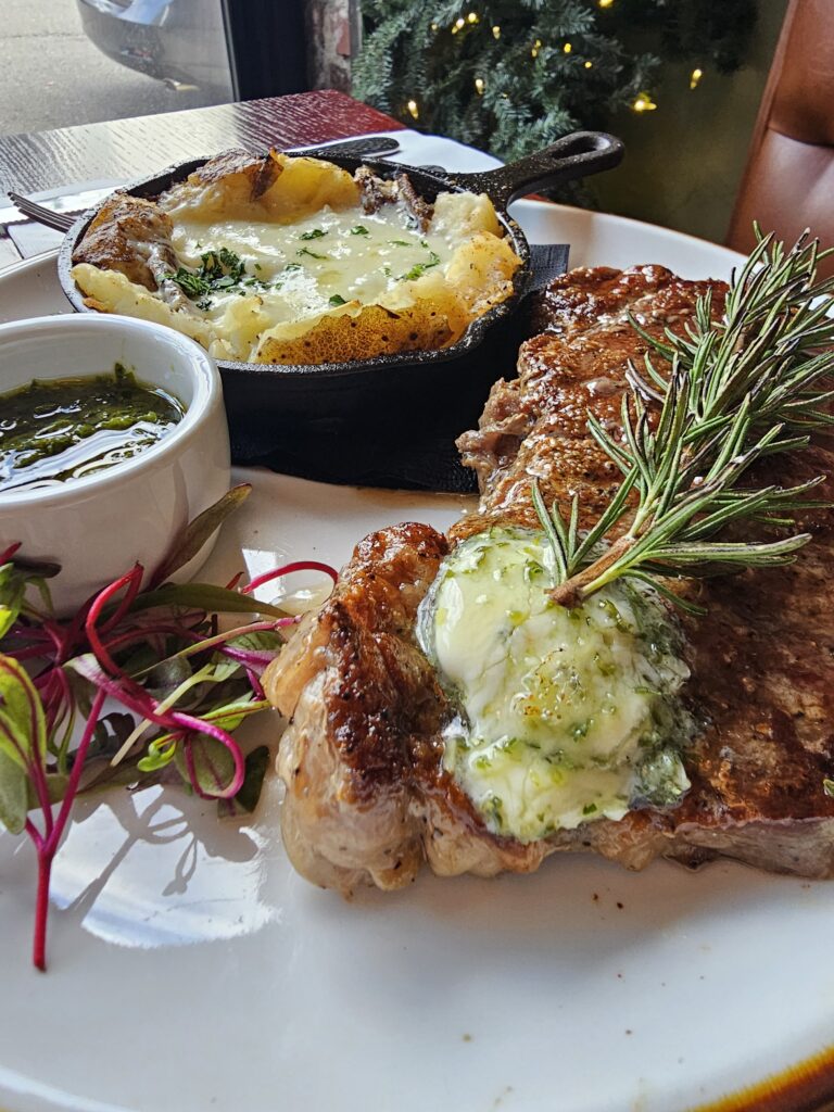 Thick seared steak with melting herb butter and rosemary at Camp West beside a bubbling cast iron potato gratin and green sauce